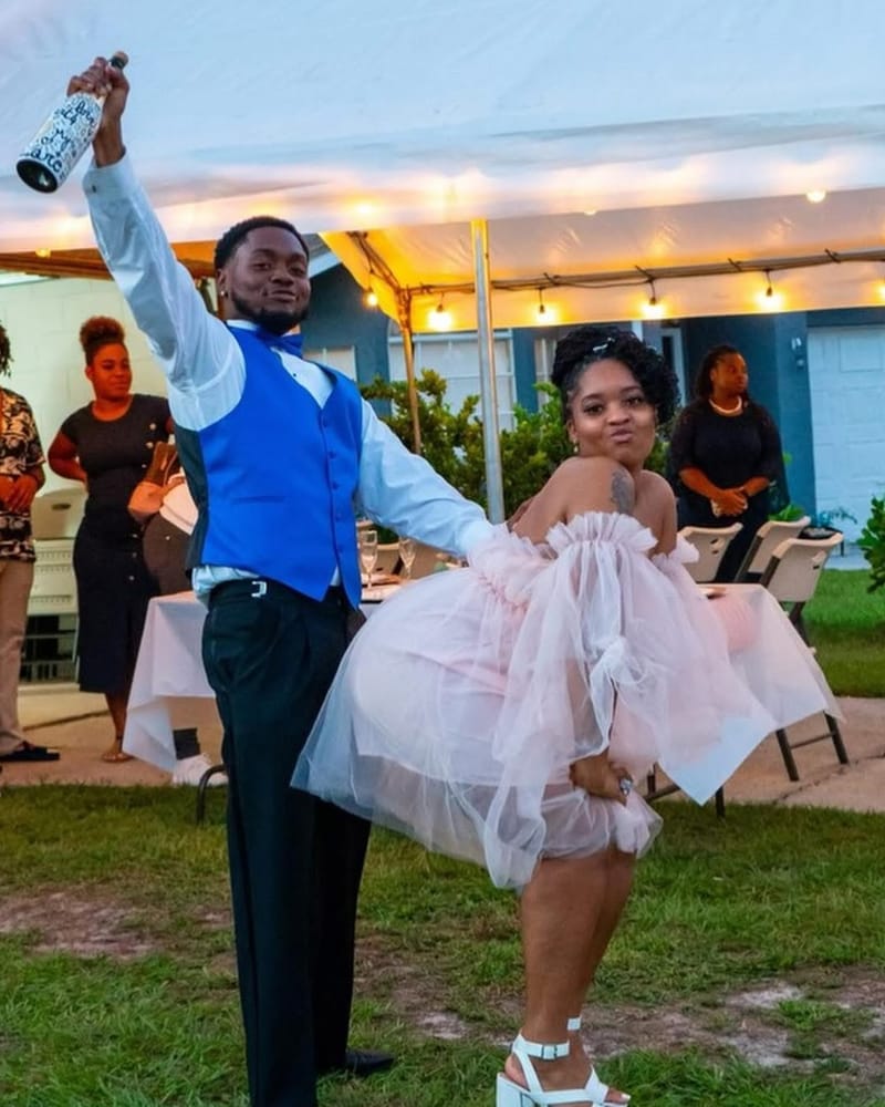 Trent now, in a blue vest, holding a bottle high at a celebration, a woman in a pink dress beside him